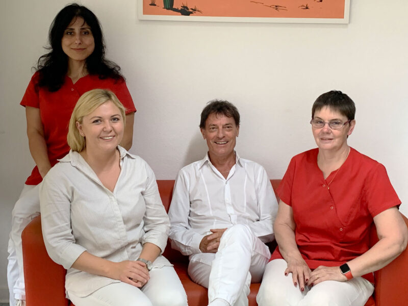 Vier lächelnde Personen auf einem roten Sofa. 3 Frauen und ein Mann. Die Ärztin und der Arzt in weißem Hemd. Die beiden Arzthelferinnen in rotem Hemd.